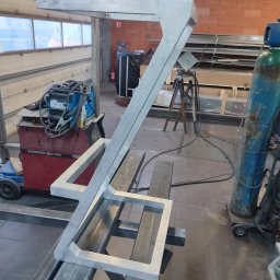 BAJ-DESIGN Błażej Dostatni - Szara, metalowa konstrukcja w kształcie litery 'Z' z kwadratową podstawą, leżąca na metalowych profilach w warsztacie, widoczny spawacz, butla z gazem i okno.