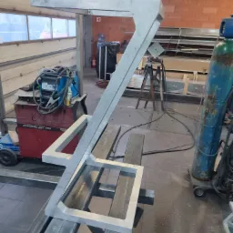 Szara, metalowa konstrukcja w kształcie litery 'Z' z kwadratową podstawą, leżąca na metalowych profilach w warsztacie, widoczny spawacz, butla z gazem i okno.