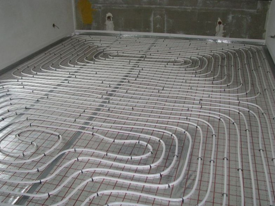 Instalacja ogrzewania podłogowego: białe rury ułożone w pętle na siatce z czerwonymi kwadratami, widoczne surowe ściany w tle.