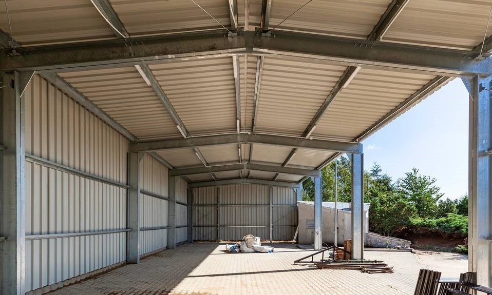 Wnętrze hali magazynowej z metalową konstrukcją, widoczne ściany z blachy falistej, betonowa podłoga z widocznymi plamami i workami w tle, częściowo zadaszona konstrukcja.