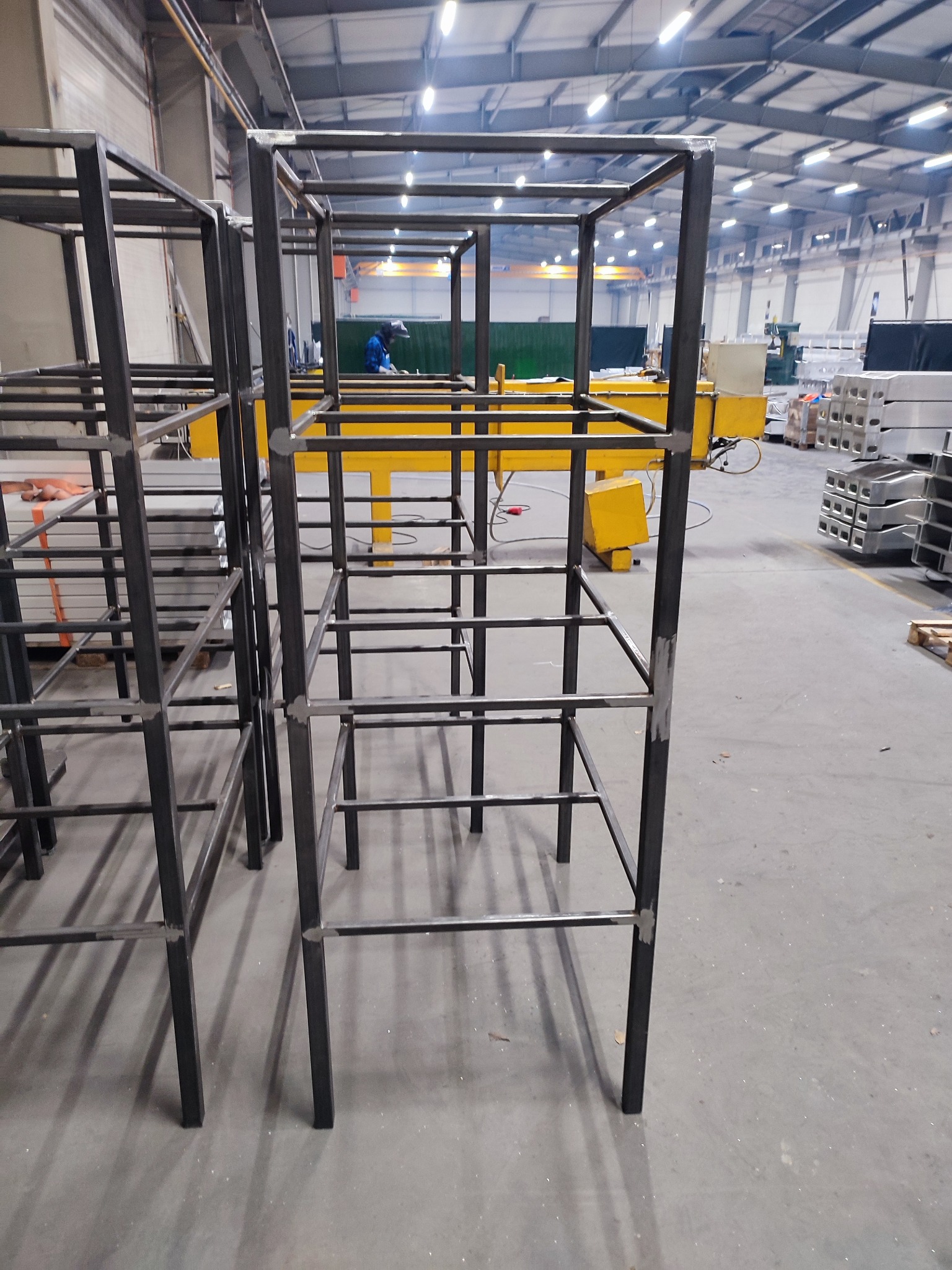 Metalowe szkielety konstrukcji spawane w hali produkcyjnej, widoczna osoba w kombinezonie spawalniczym w tle.