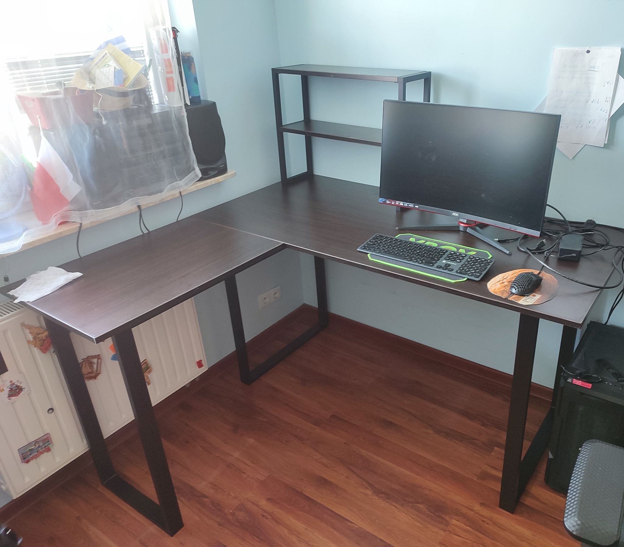 Narożne biurko z ciemnego drewna z metalowymi nogami, monitor, klawiatura i mysz na blacie, dodatkowa półka nad biurkiem, widok z góry na podłogę z paneli.