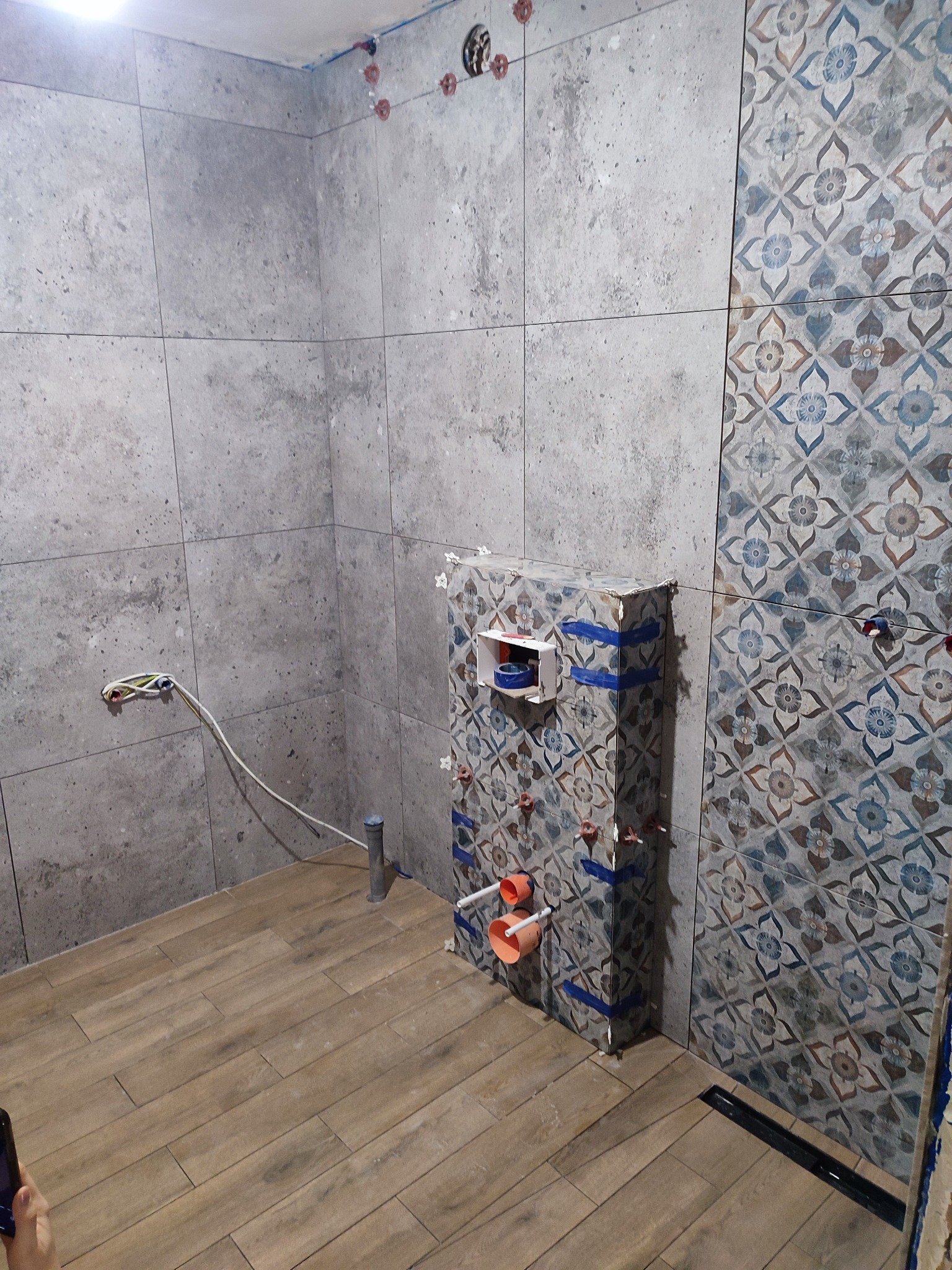 Wnętrze łazienki w trakcie remontu: położone płytki imitujące drewno na podłodze, częściowo wyłożone płytki na ścianach, w tym jedna ściana z wzorem kwiatowym, widoczne elementy instalacji...