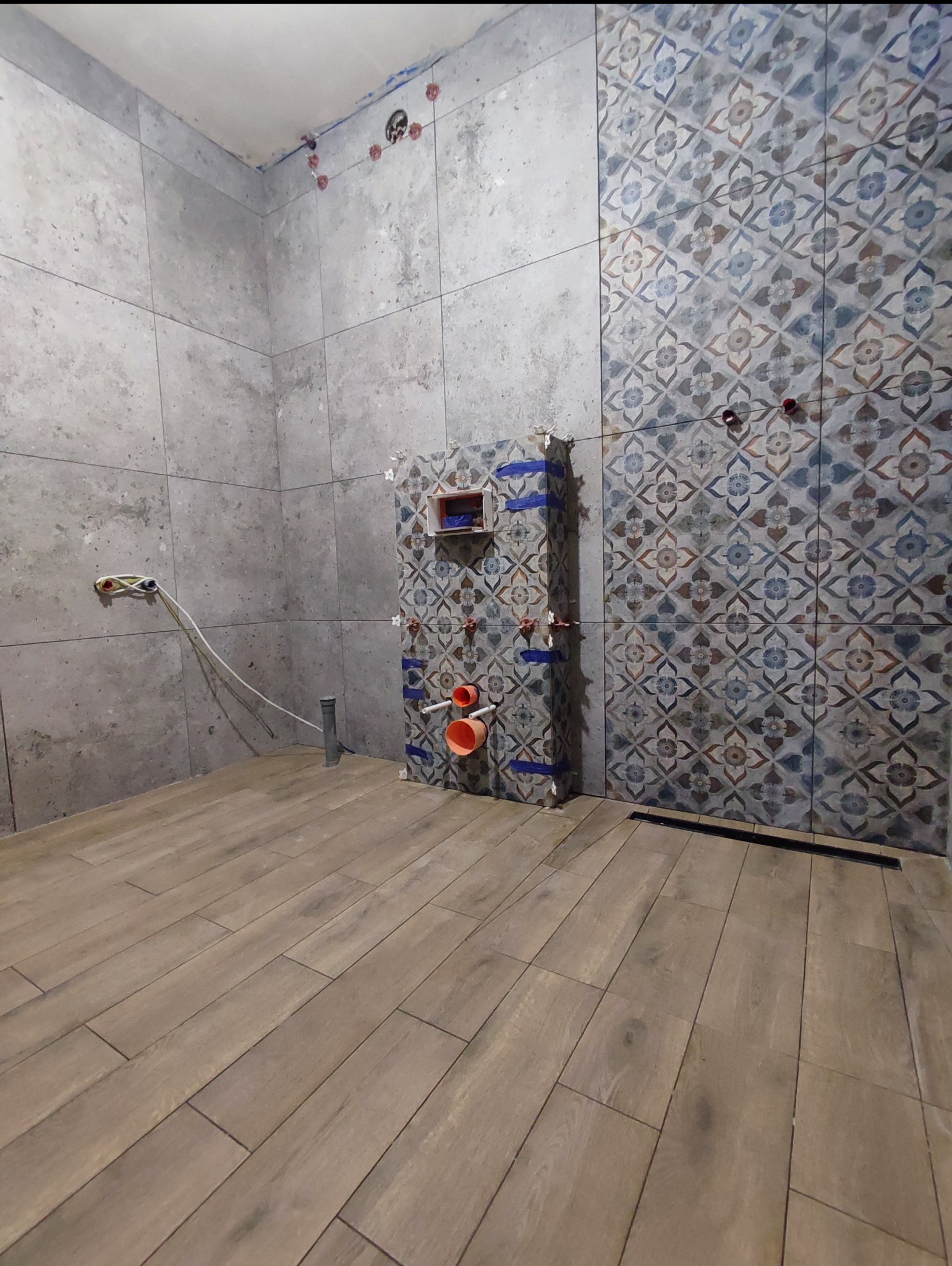 Wnętrze łazienki w trakcie remontu: częściowo ułożone płytki ścienne w dwóch wzorach, widoczne instalacje hydrauliczne i elektryczne, drewnopodobna podłoga.