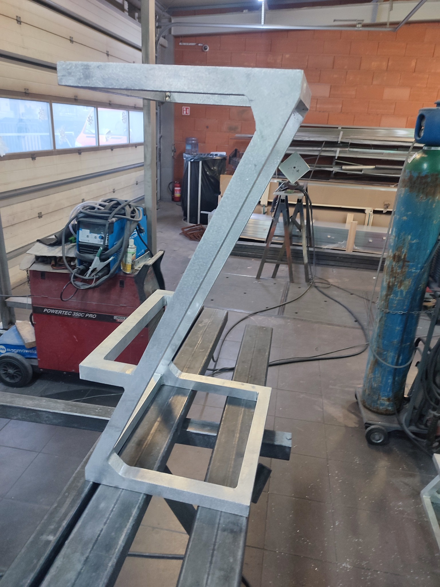 Szara, metalowa konstrukcja w kształcie litery 'Z' z kwadratową podstawą, leżąca na metalowych profilach w warsztacie, widoczny spawacz, butla z gazem i okno.