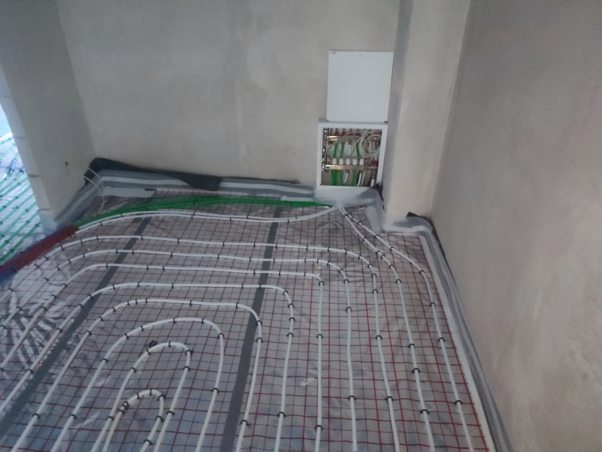 Rozłożona instalacja ogrzewania podłogowego z rurkami na siatce, widoczna szafka rozdzielcza z zielonymi i białymi przewodami na ścianie w surowym wnętrzu.
