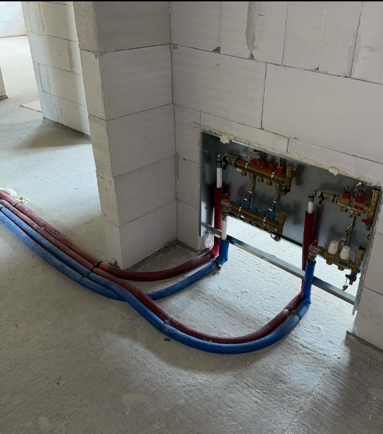 Rozdzielacz ogrzewania podłogowego w metalowej szafce w ścianie z betonu komórkowego, rury doprowadzające wodę w kolorze czerwonym i niebieskim, surowy beton na podłodze.