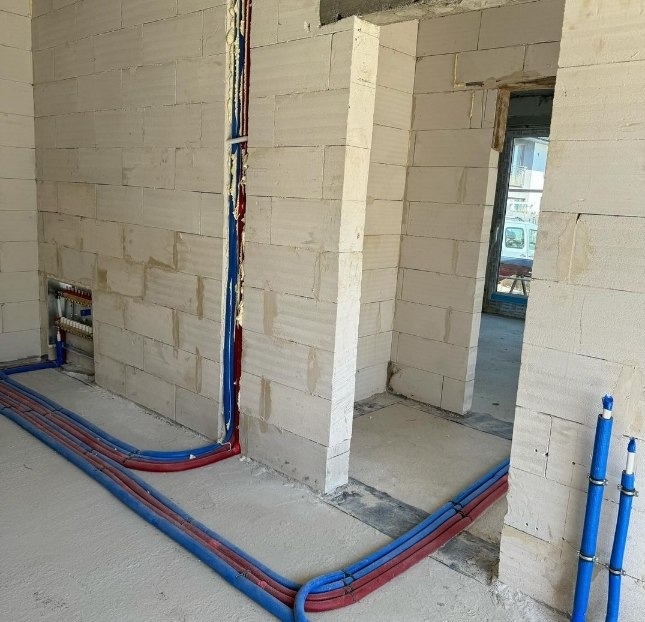 Wnętrze w trakcie budowy z widocznymi instalacjami wodno-kanalizacyjnymi prowadzonymi po podłodze i w ścianach z białego bloczka.
