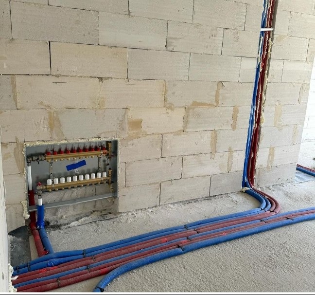 Rozdzielacz ogrzewania podłogowego zamontowany w ścianie z bloczków betonowych, z widocznymi rurami doprowadzającymi w kolorze niebieskim i czerwonym, zabezpieczonymi pianką montażową.