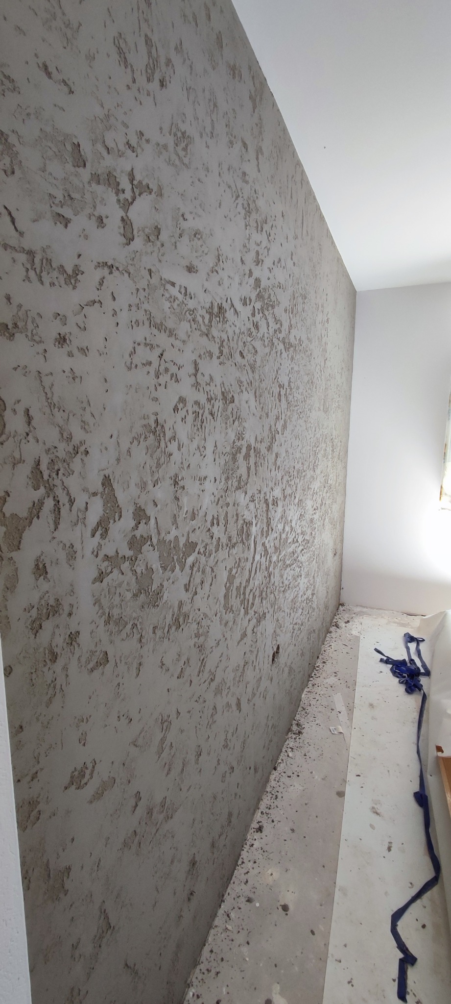Ściana wewnętrzna z dekoracyjną, szarą fakturą, widoczne drobne nierówności i fragment zabezpieczonej podłogi po malowaniu.