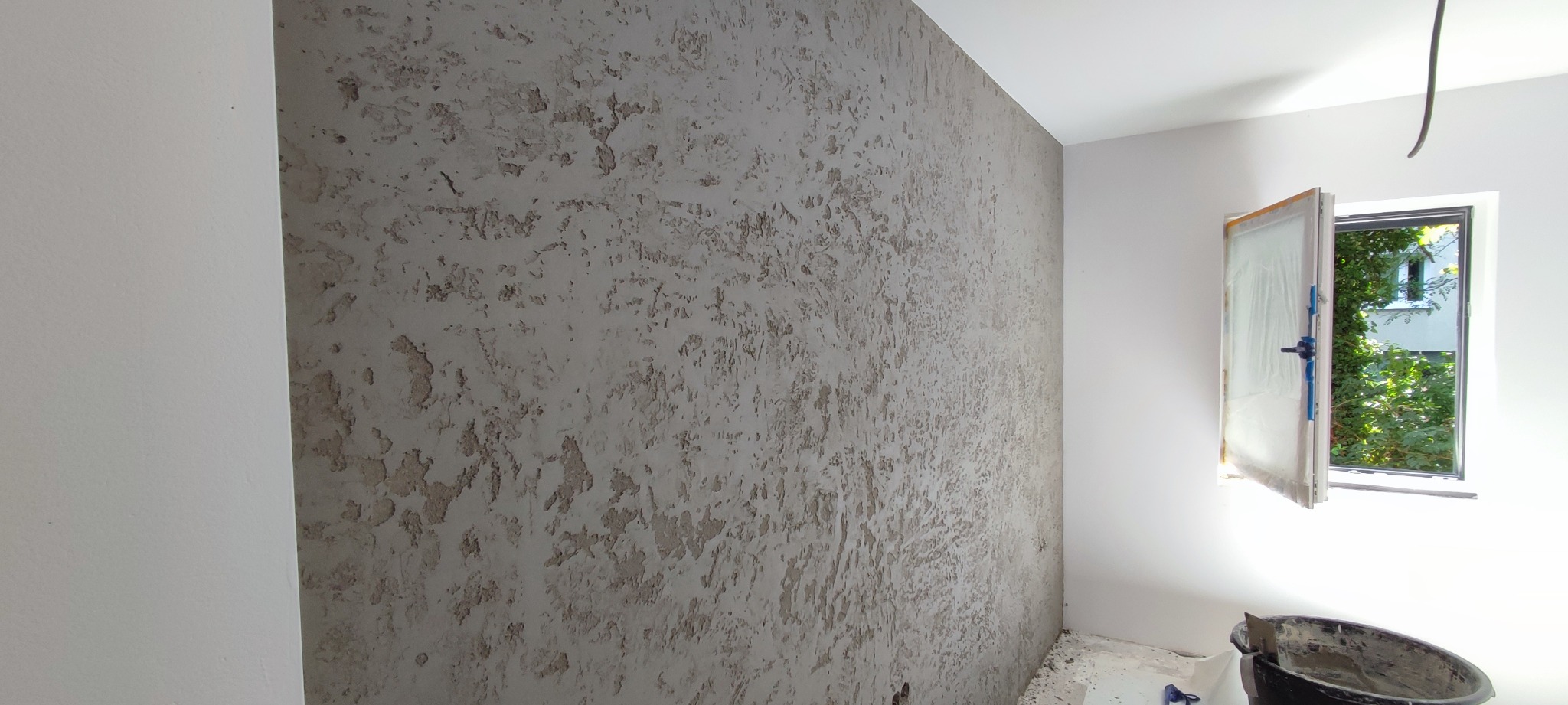 Pokój po nałożeniu dekoracyjnej struktury na ścianę, widoczne otwarte okno z widokiem na zieleń, wiadro i narzędzia malarskie w rogu.