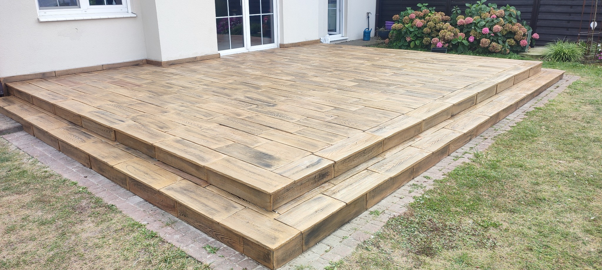 Betonowy taras z dwoma stopniami, imitujący drewniane deski, obrzeże z kostki brukowej, trawnik i hortensje w tle.