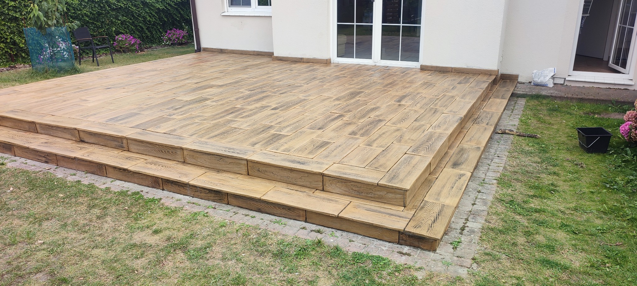 Wykończony taras z betonu stemplowanego imitującego drewno, z dwustopniowymi schodami, widok z poziomu trawnika, w tle ściana budynku z białymi drzwiami balkonowymi.