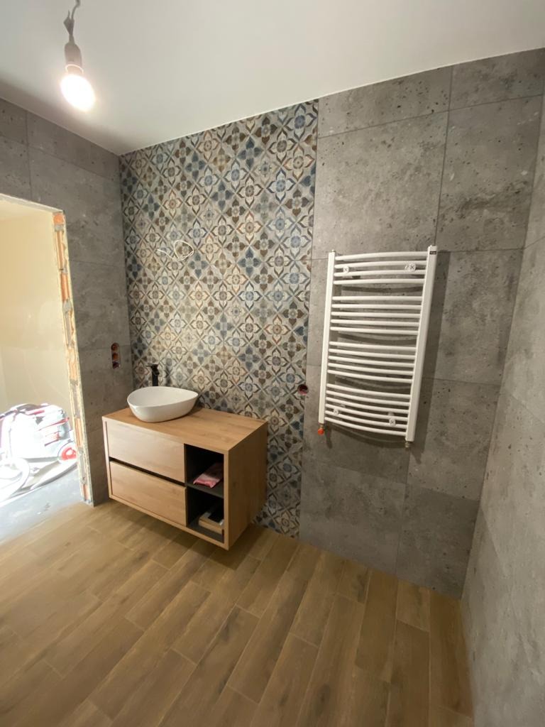 Wnętrze łazienki w trakcie remontu: drewnopodobna podłoga, umywalka nablatowa na drewnianej szafce z szufladami, dekoracyjna mozaika na ścianie oraz grzejnik drabinkowy na szarej płytce. W tle...
