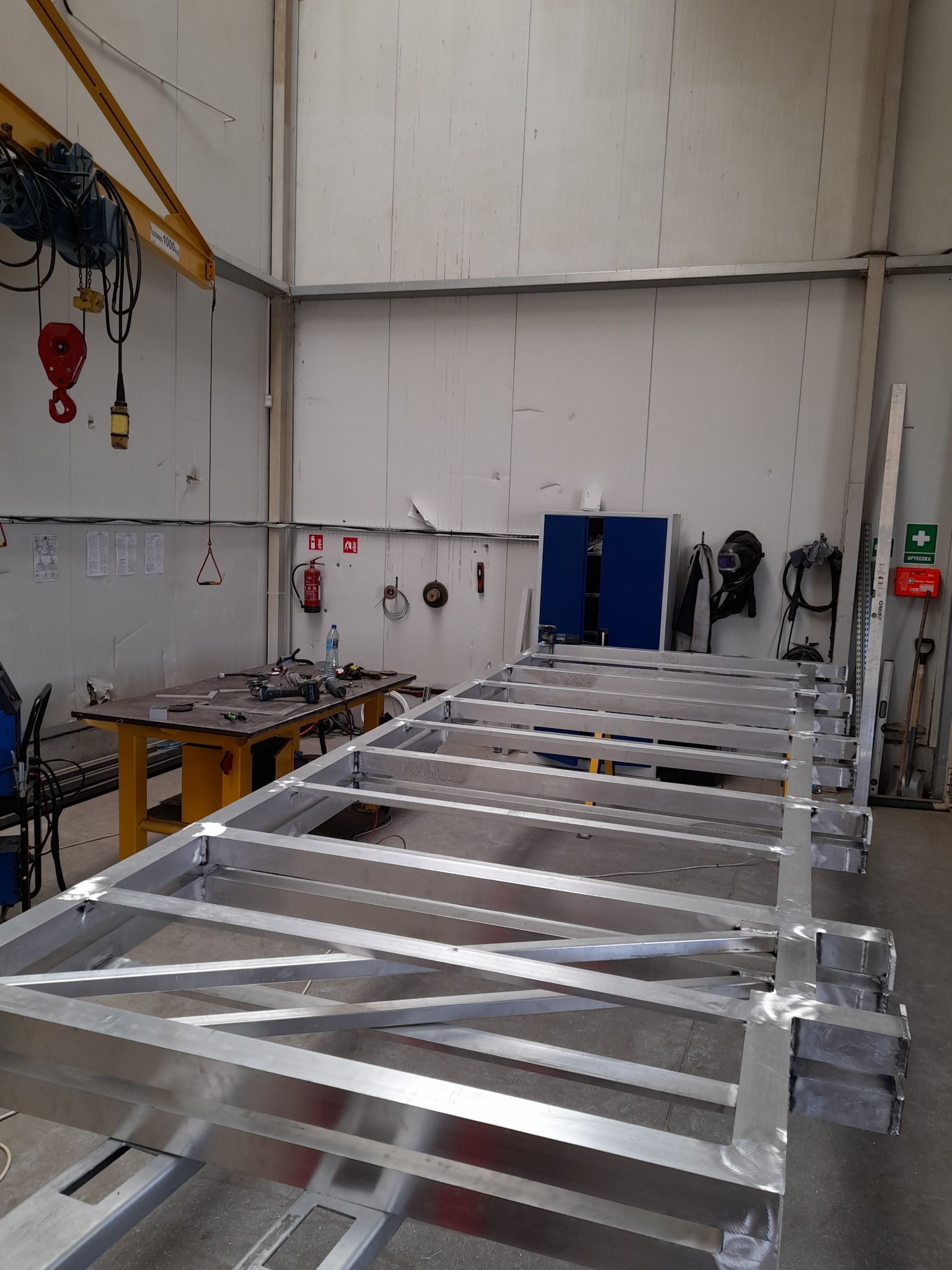 Metalowa konstrukcja ramy, prawdopodobnie podestu lub platformy, z widocznymi spawami, w hali produkcyjnej, z narzędziami na stole w tle i suwnicą pod sufitem.