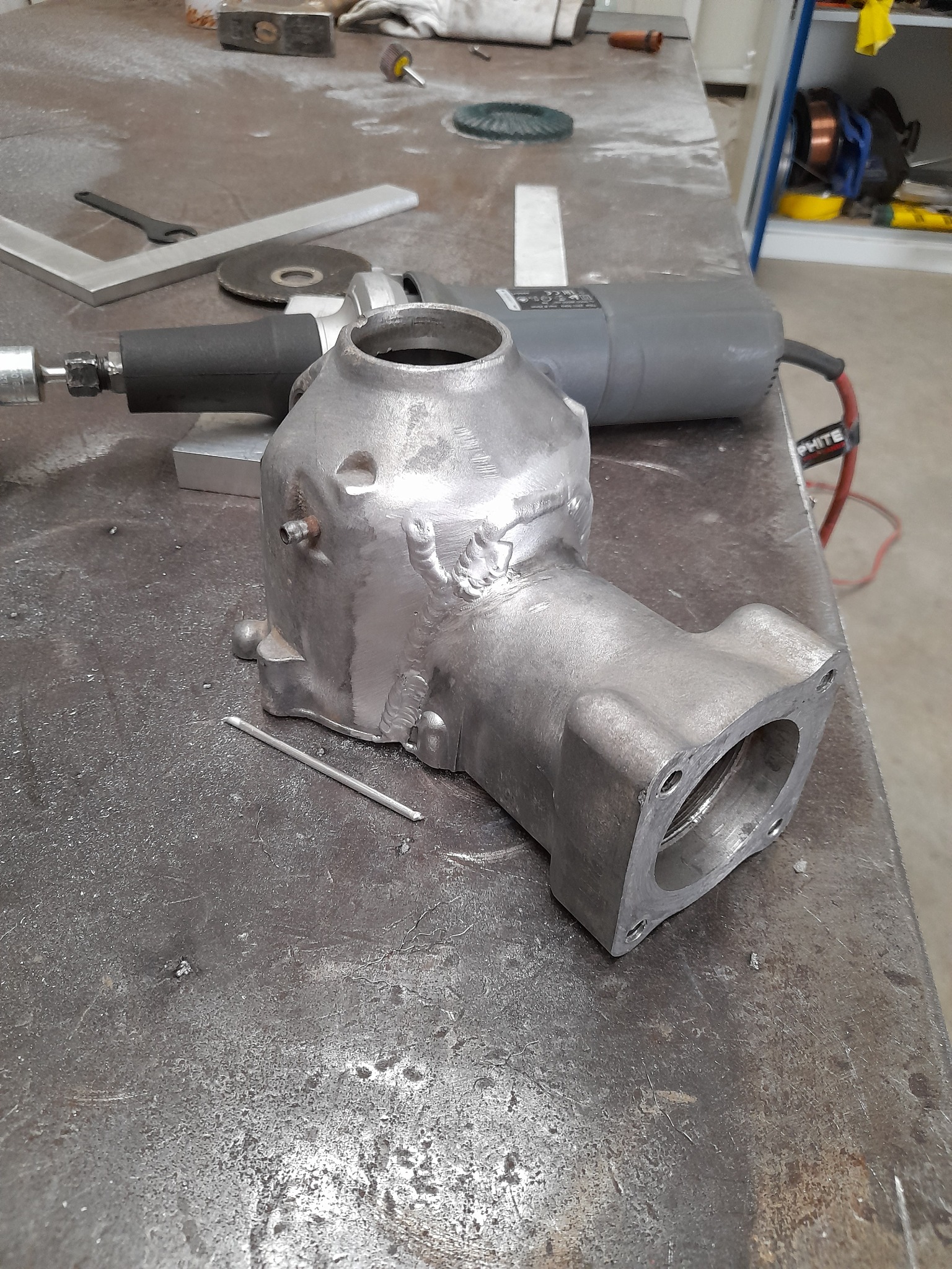 Surowy odlew metalowy z widocznymi śladami po spawaniu i obróbce szlifierskiej, leżący na warsztatowym stole obok szlifierki kątowej i innych narzędzi.