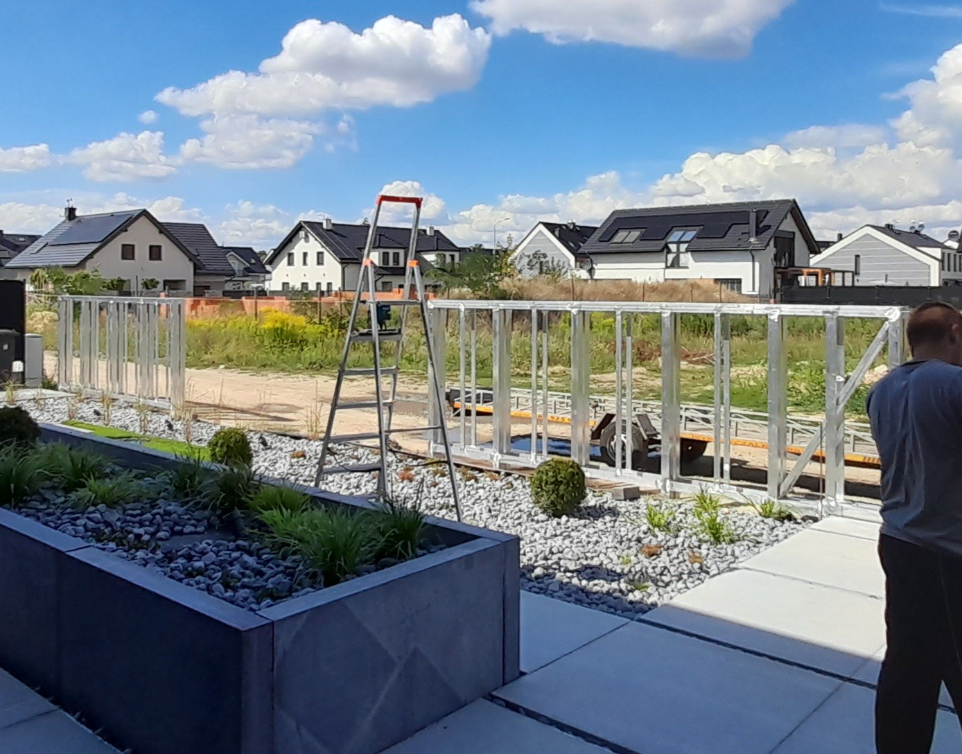 Montaż aluminiowego ogrodzenia panelowego na tle zabudowy jednorodzinnej, widoczna drabina i przyczepka z elementami ogrodzenia.