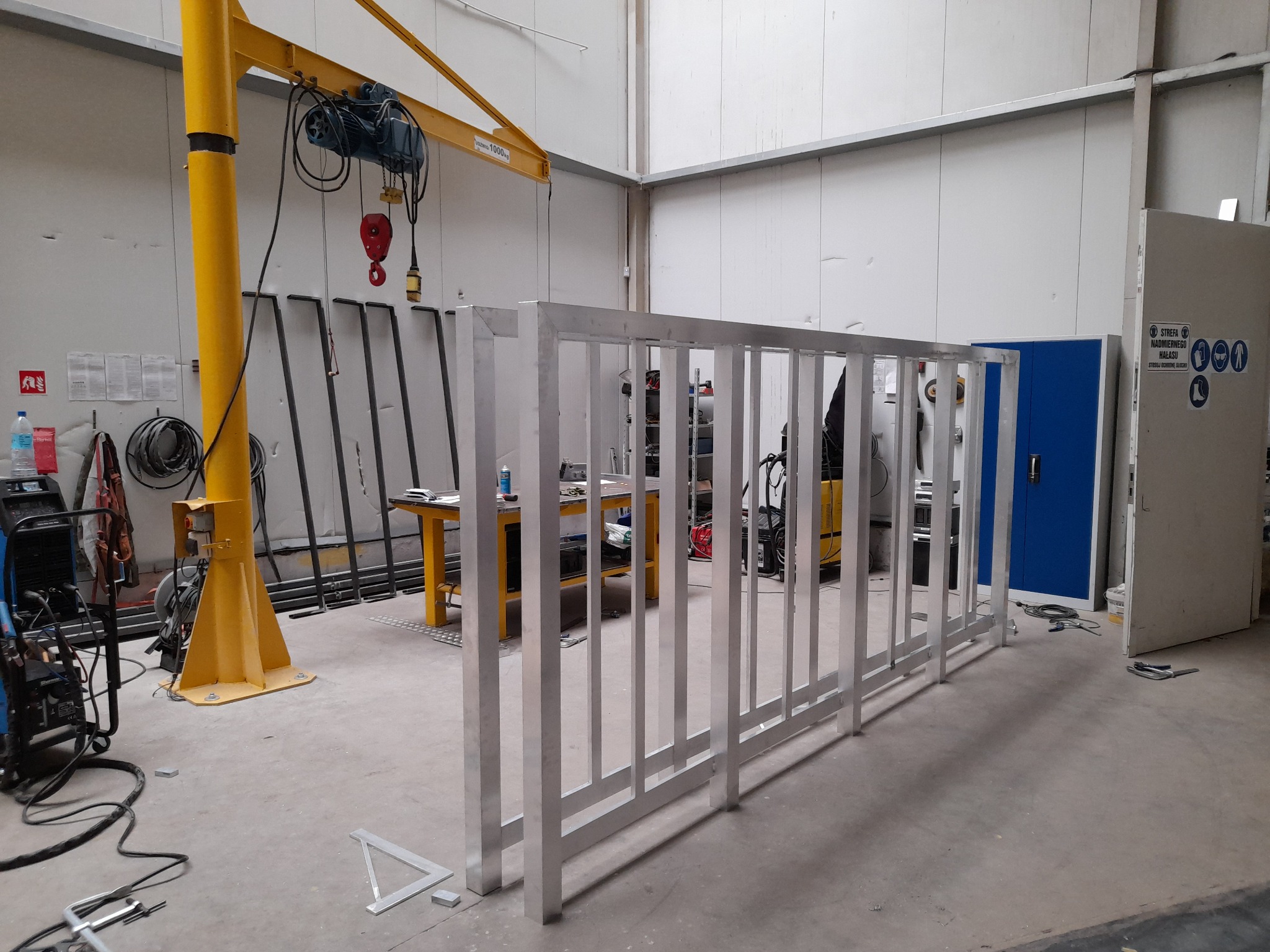 Wnętrze hali produkcyjnej z surową, aluminiową konstrukcją ogrodzenia lub balustrady, widoczny żółty suwnicowy dźwig, spawarka i narzędzia, w tle metalowe profile i stół warsztatowy.