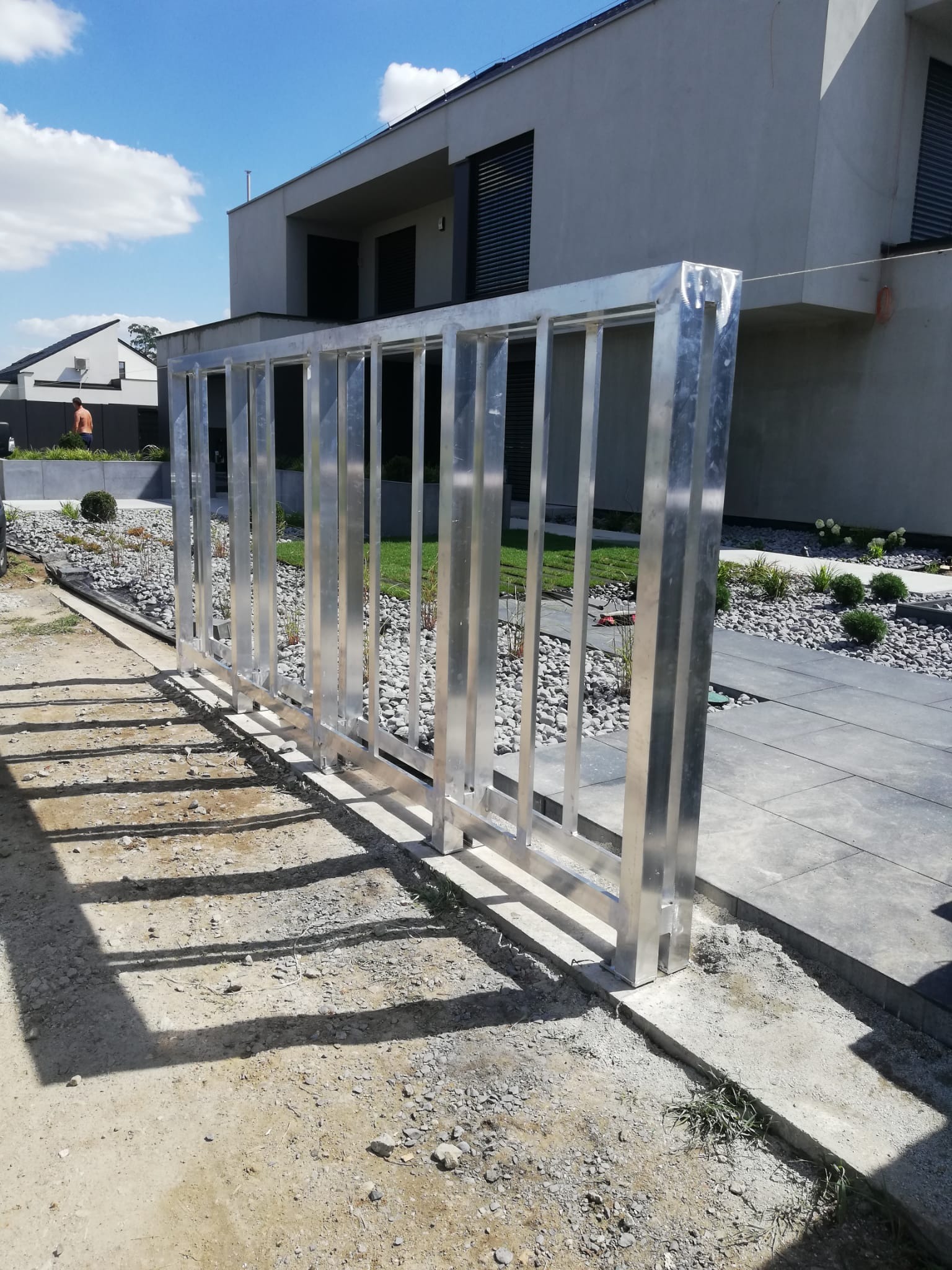 Surowa, metalowa konstrukcja bramy przesuwnej w trakcie montażu, widoczne betonowe fundamenty i niezabezpieczone podłoże, nowoczesny dom w tle.