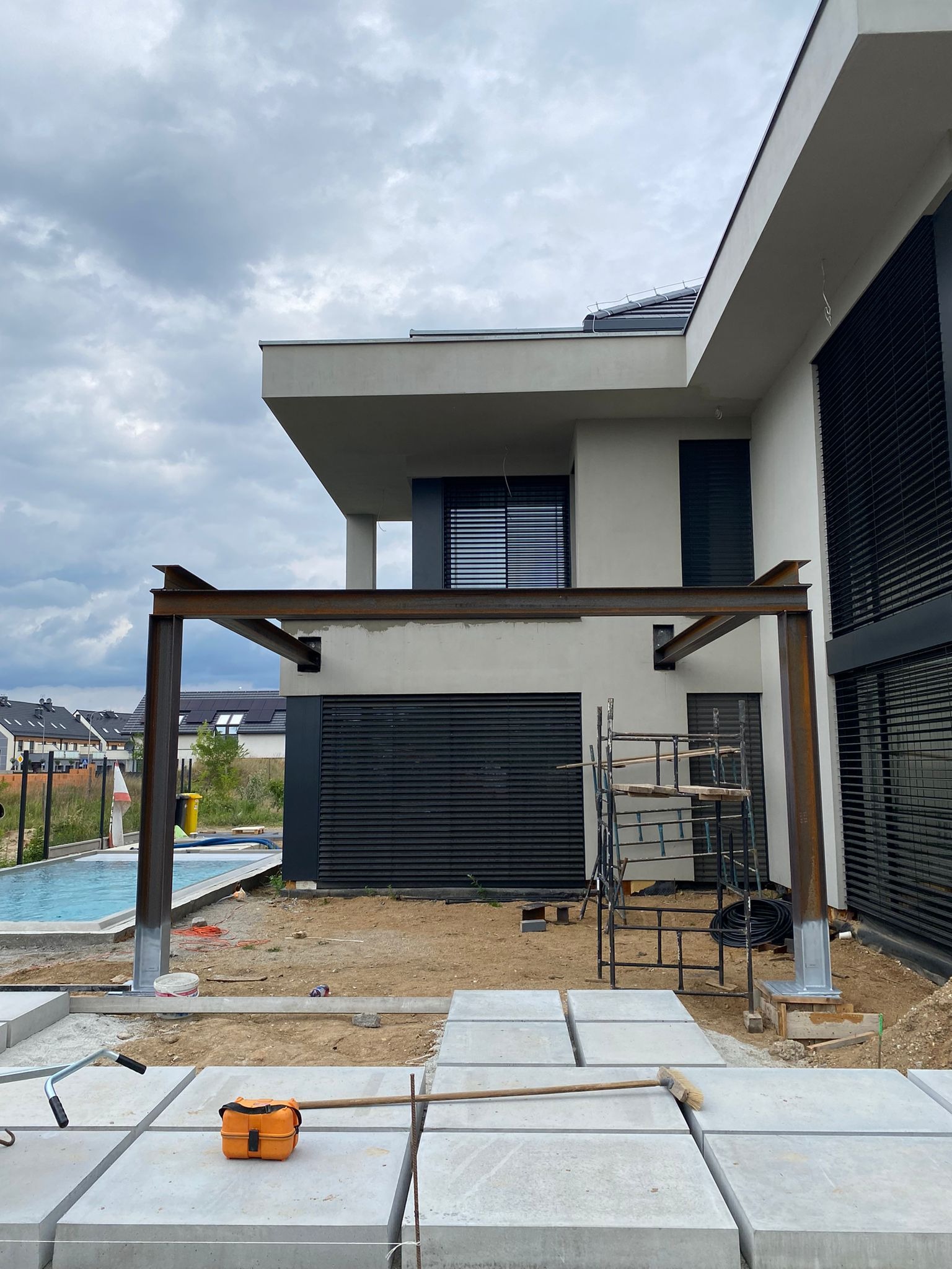 Montaż metalowej konstrukcji z profili stalowych przy nowoczesnym domu z basenem, widoczne bloczki betonowe, rusztowanie i narzędzia.