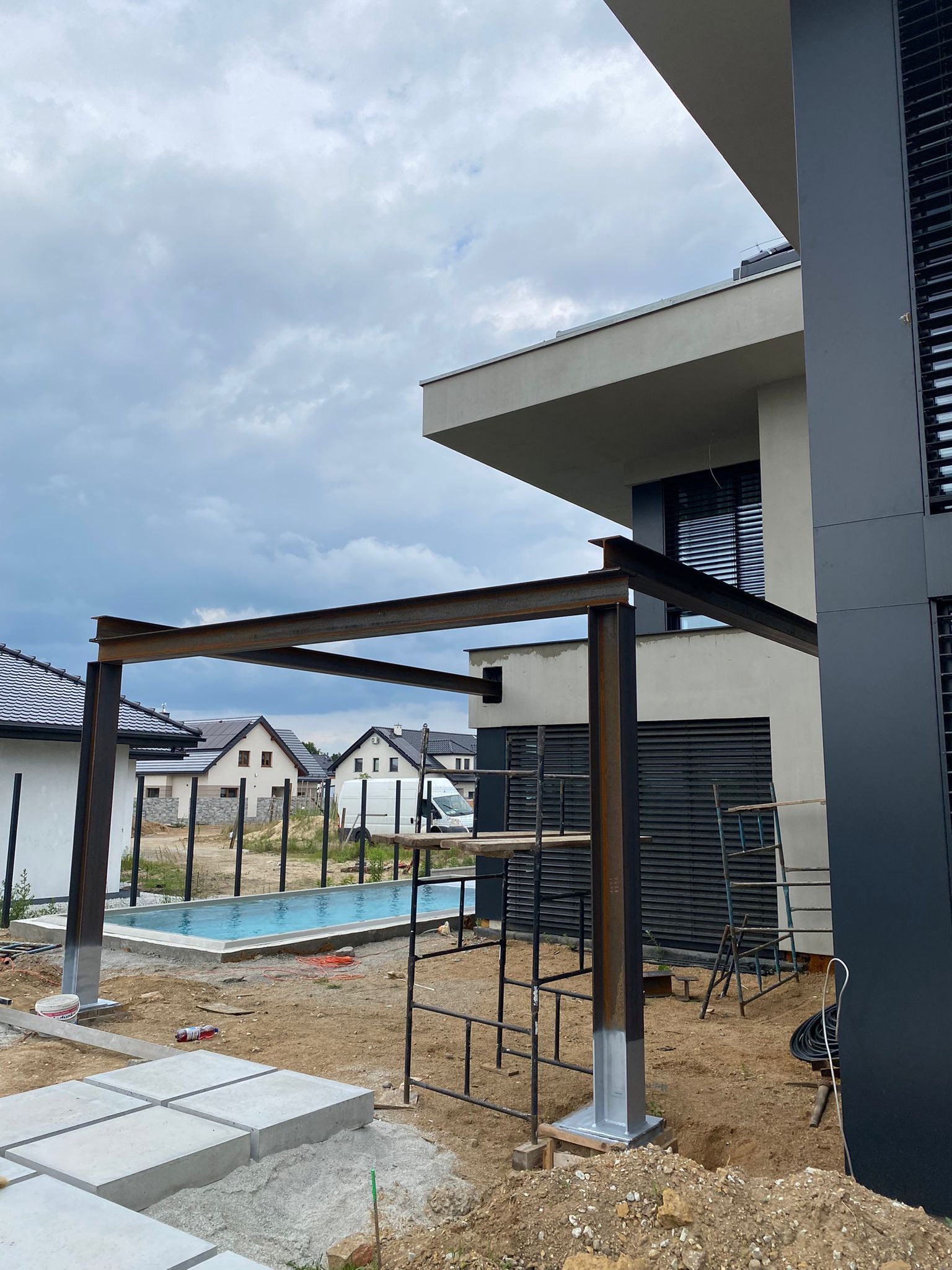 Konstrukcja metalowa z profili stalowych w trakcie montażu na zewnątrz nowoczesnego domu, widoczny basen i rusztowania, ziemia wokół w stanie budowy.