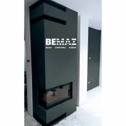 Nowoczesny, grafitowy kominek narożny z szybą, z logo firmy BEMAZ, prezentujący bramy, ogrodzenia i kominki, w surowym wnętrzu w trakcie wykończenia.