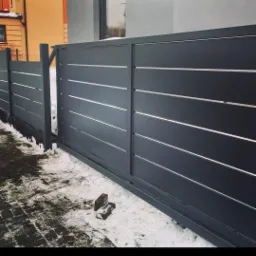 Nowoczesna, przesuwna brama w kolorze antracytu z poziomymi, srebrnymi akcentami, częściowo zasypana śniegiem, z widocznymi kostkami brukowymi.