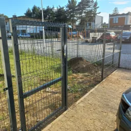 Otwarta furtka w panelowym ogrodzeniu posesji w Jaśkowicach, widok na trawnik i zaparkowane samochody za ogrodzeniem.