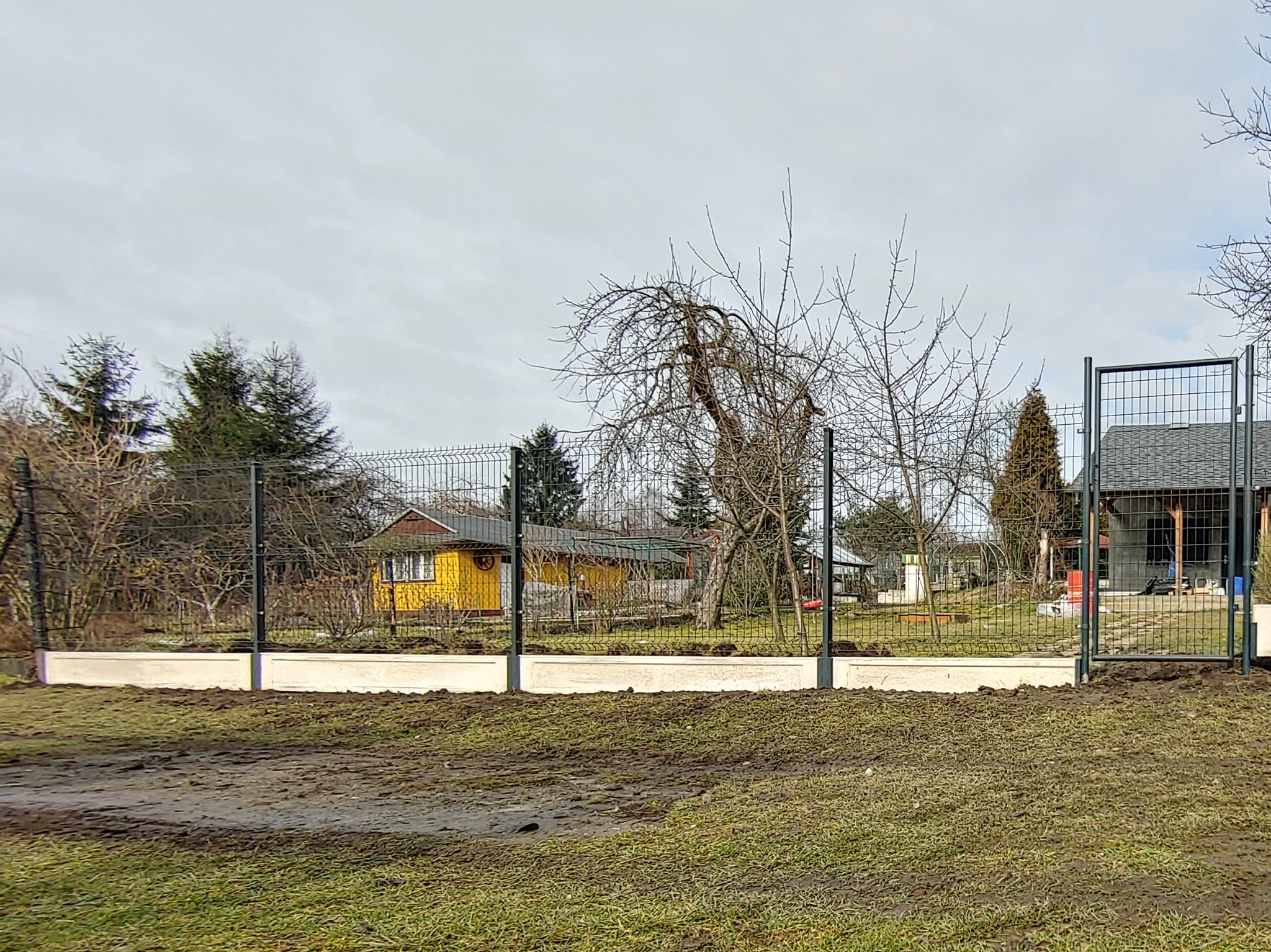 Nowoczesne ogrodzenie panelowe z bramą wjazdową, osadzone na betonowej podmurówce, widok na ogród z domkami letniskowymi w tle.