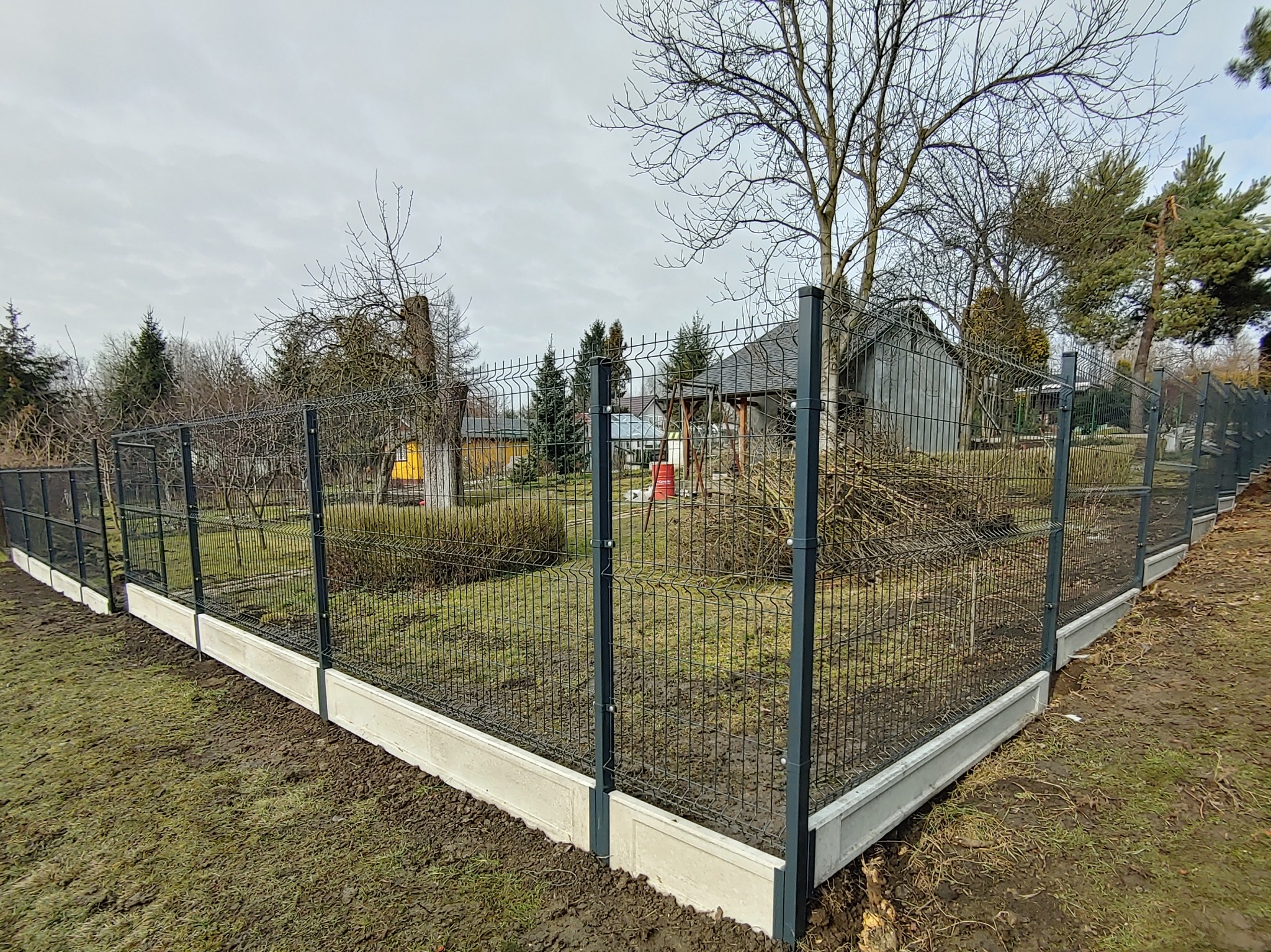 Nowo zamontowane ogrodzenie panelowe z podmurówką betonową, widoczne słupki i siatka, otoczenie posesji z drzewami i budynkami w tle, pochmurny dzień.