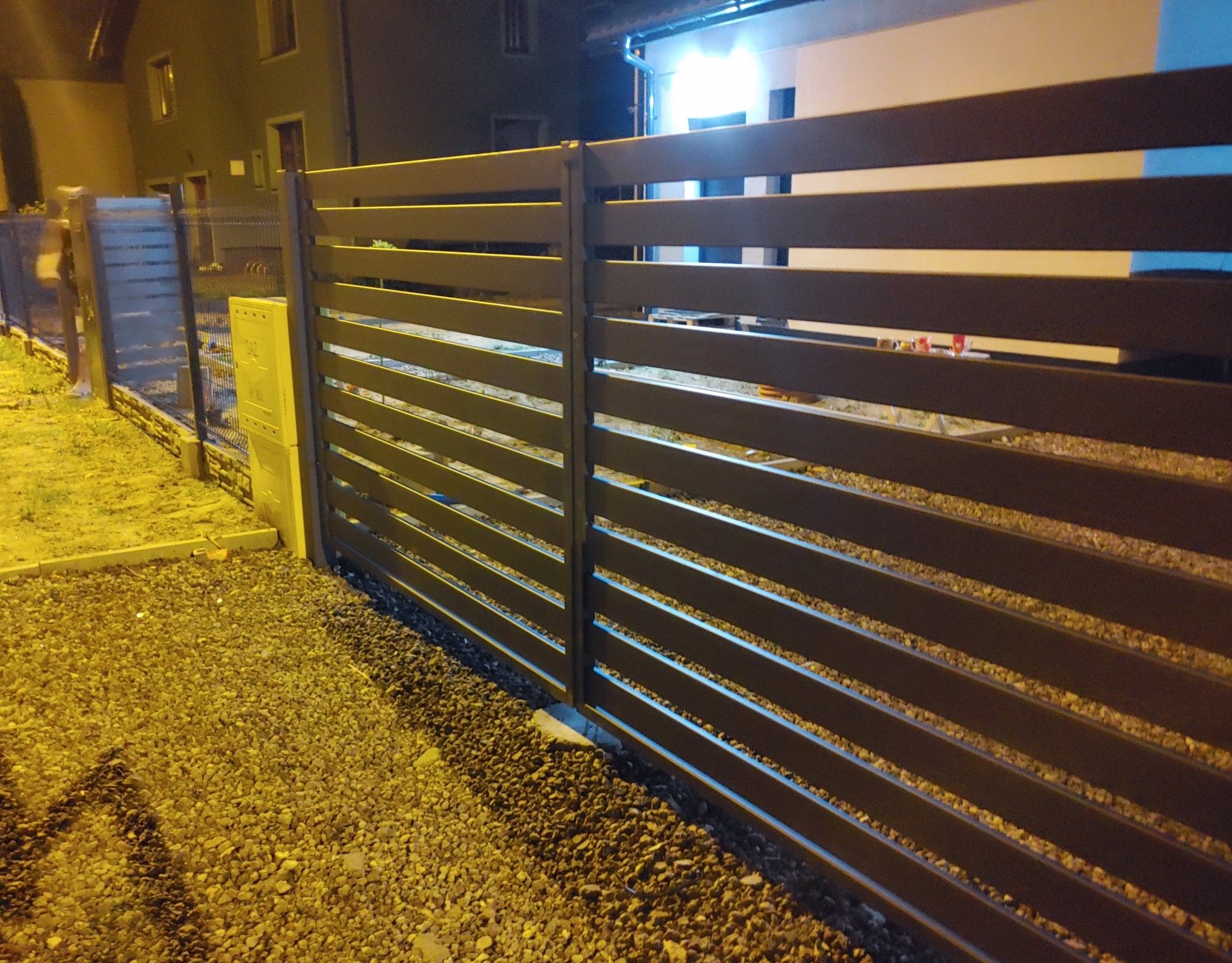 Nowoczesne, poziome przęsła ogrodzeniowe w kolorze antracytowym, widok z bliska na tle oświetlonego budynku mieszkalnego, z widoczną skrzynką gazową i fragmentem ogrodzenia panelowego w tle.