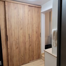 PU Sawa - Wnętrze: drewniana szafa przesuwna z czarnymi uchwytami, jasne panele podłogowe, biała ściana i oświetlenie sufitowe. Widok zza futryny.