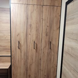 PU Sawa - Drewniana szafa wnękowa z czarnymi uchwytami, dopasowana do podłogi i ściany. Minimalistyczny design, widoczny fragment szafki nocnej i drzwi.