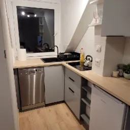 Nowoczesna, jasna kuchnia w szarościach z drewnianym blatem. Widoczne AGD, czarny zlew i płyta grzewcza, oświetlenie punktowe i drewniana podłoga.