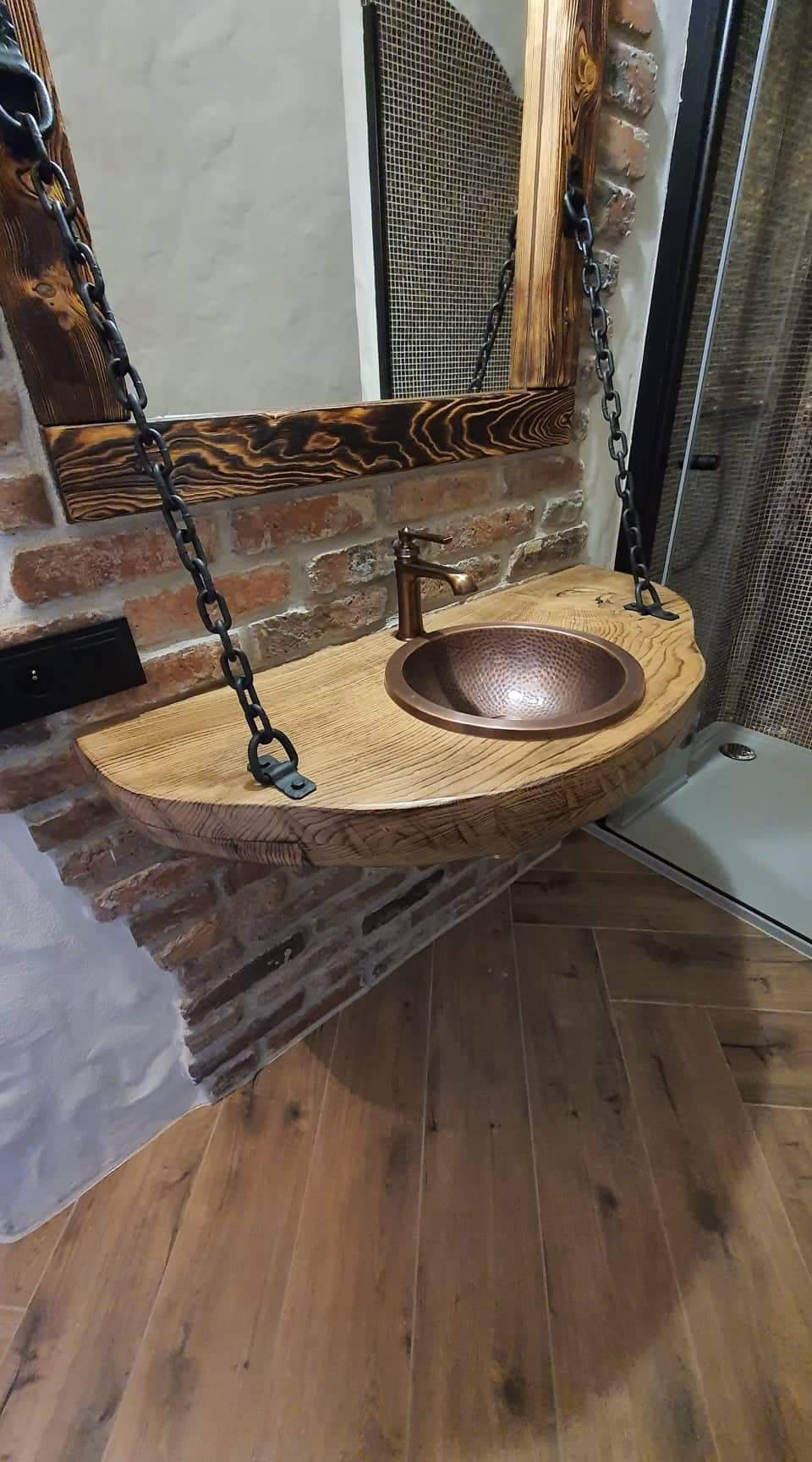 Rustykalna umywalka z miedzianą misą i drewnianym blatem zawieszona na łańcuchach, na tle ściany z cegły i kabiny prysznicowej.