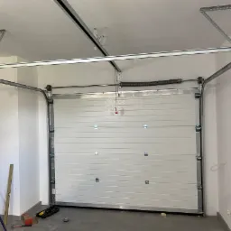 Montaż segmentowej bramy garażowej z widocznymi prowadnicami, sprężyną i narzędziami na podłodze w jasnym garażu.