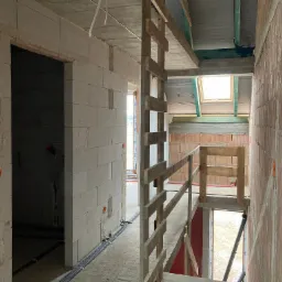 Wnętrze w stanie surowym: ściany z bloczków, widoczne kable elektryczne w peszlach, drewniana konstrukcja schodów i okno dachowe.