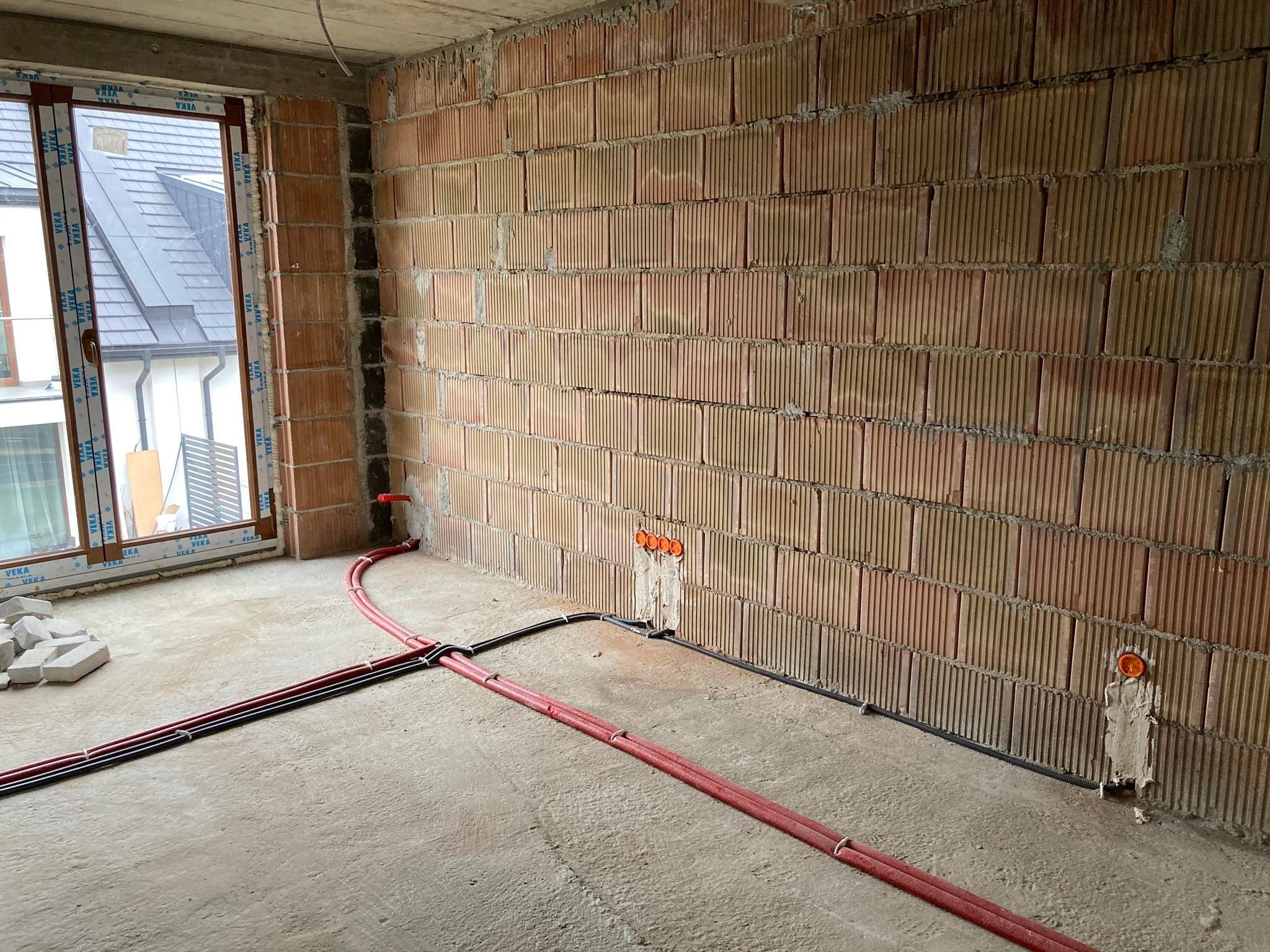Wnętrze surowego budynku w trakcie prac, widoczne czerwone i czarne rury instalacji elektrycznej prowadzone po podłodze i ścianach z cegły ceramicznej dziurawki, puszki instalacyjne w ścianach...