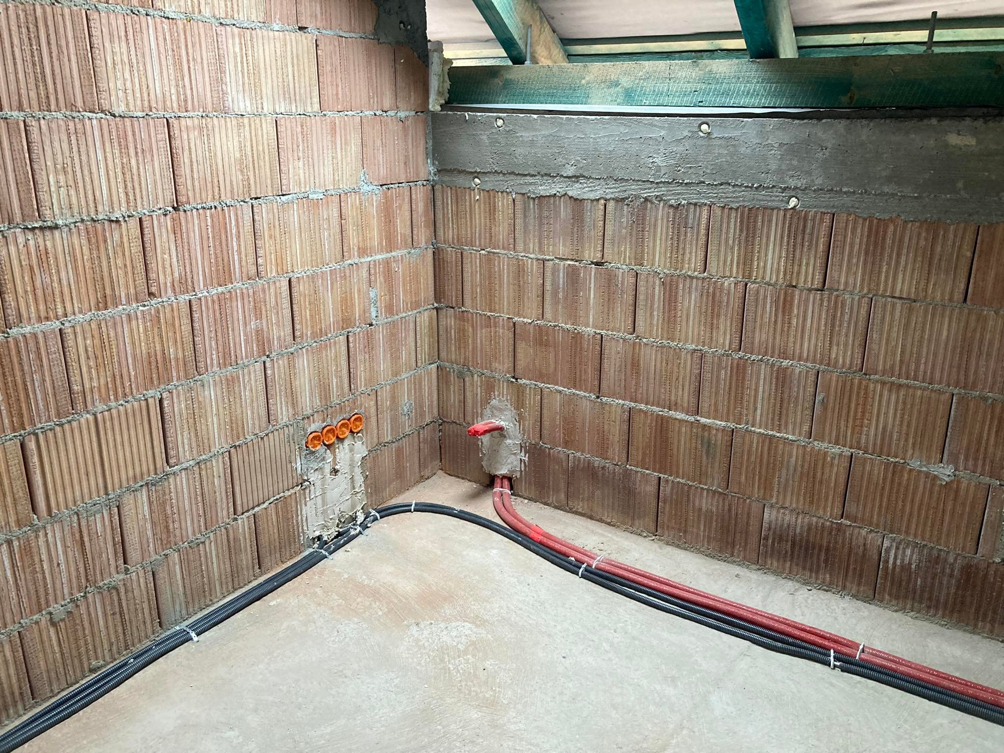 Wnętrze w trakcie budowy z widocznymi przewodami elektrycznymi w peszlach, częściowo zamurowanymi w ścianie z cegły kratówki. Widoczne pomarańczowe puszki instalacyjne pod tynkiem oraz surowy strop...