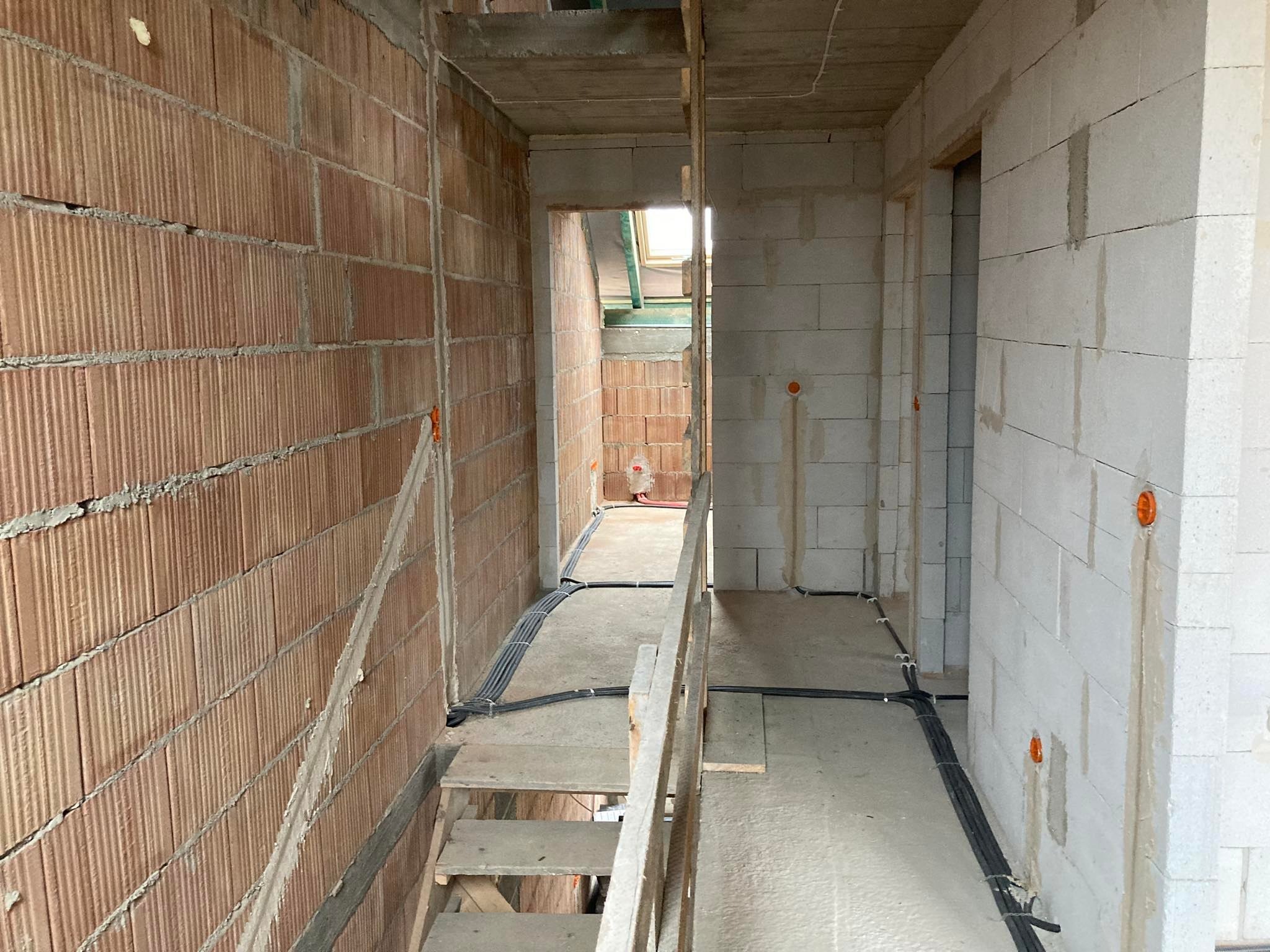 Wnętrze surowego budynku w trakcie budowy, widoczne kable elektryczne prowadzone po podłodze i ścianach, otwory pod gniazdka, schody tymczasowe.
