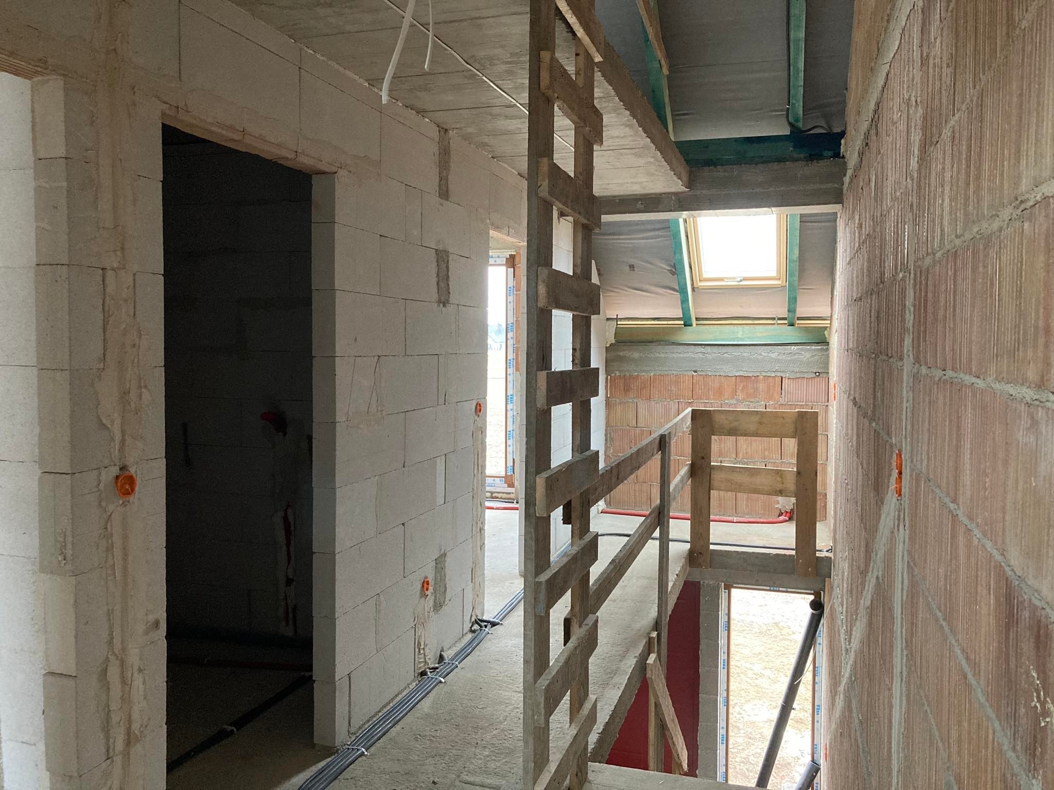 Wnętrze w stanie surowym: ściany z bloczków, widoczne kable elektryczne w peszlach, drewniana konstrukcja schodów i okno dachowe.