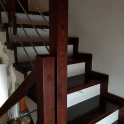 Ciemne, lakierowane stopnie z drewna egzotycznego, białe podstopnice i metalowe barierki, widok z dołu na fragment biegu schodów, widoczne słoje drewna na słupku.