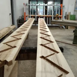 Dwie drewniane belki z wyfrezowanymi zygzakowatymi wzorami na warsztatowym stole, w tle narzędzia i maszyny stolarskie.