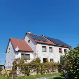 Biały dom z panelami słonecznymi na dachu z czerwoną dachówką, widok z dołu, błękitne niebo w tle, winorośl przy ścianie, drabina na trawniku.