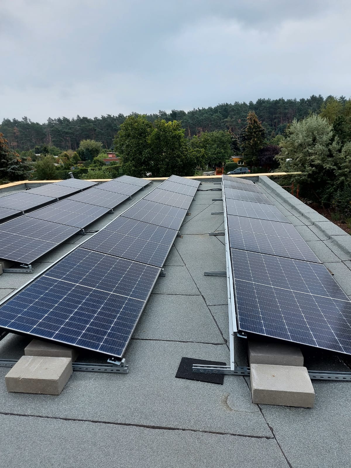 Panele słoneczne zamontowane na płaskim dachu z wykorzystaniem betonowych bloczków i metalowych konstrukcji wsporczych. W tle widoczny las i pochmurne niebo.