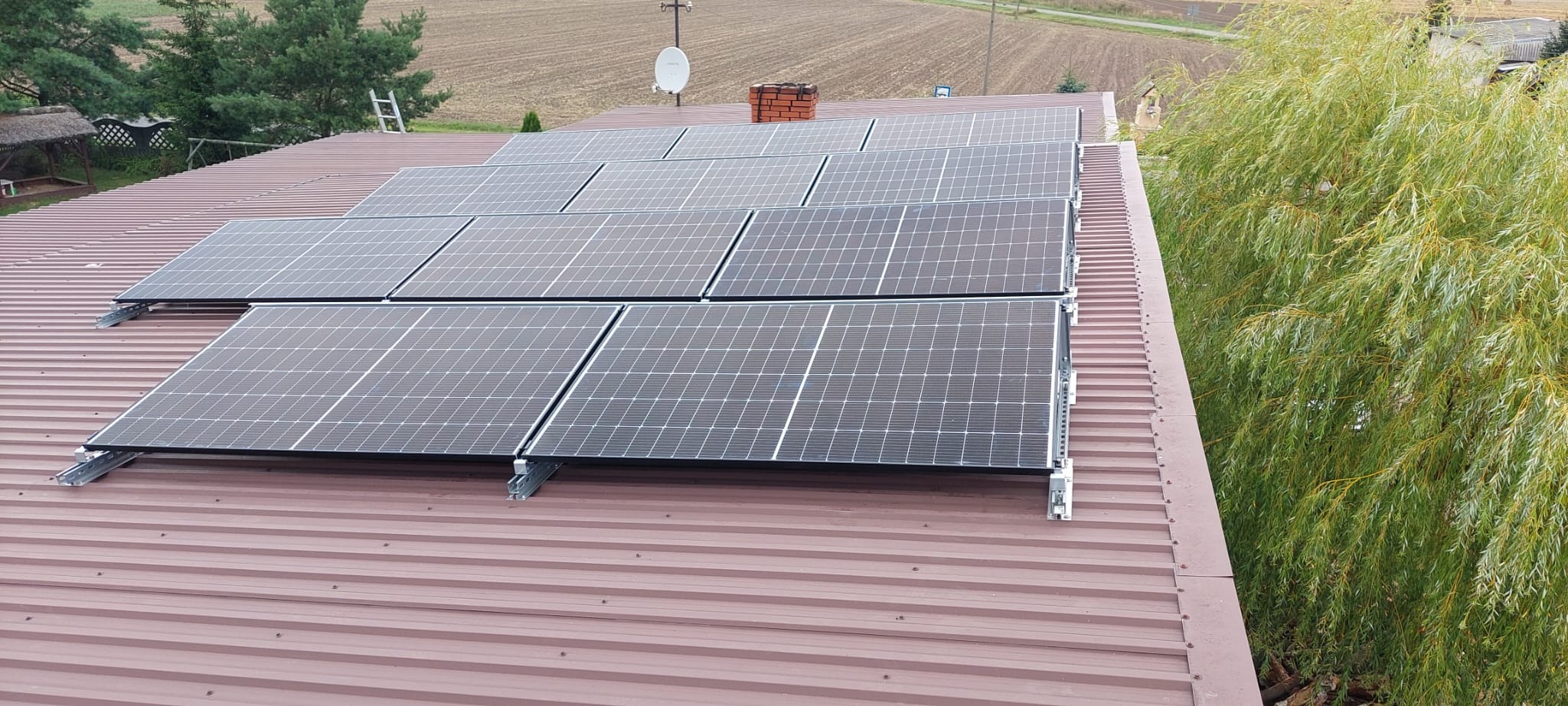 Panele słoneczne zamontowane na brązowym dachu z blachy trapezowej, widok z góry, w tle wiejski krajobraz i wierzba płacząca.