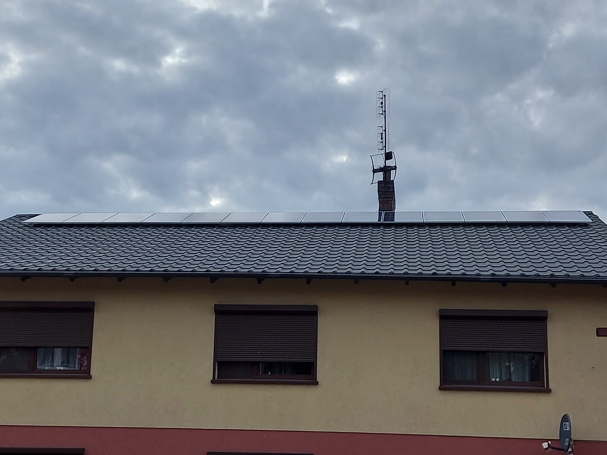 Dom z panelami słonecznymi na dachu, widoczna antena telewizyjna, pochmurne niebo.