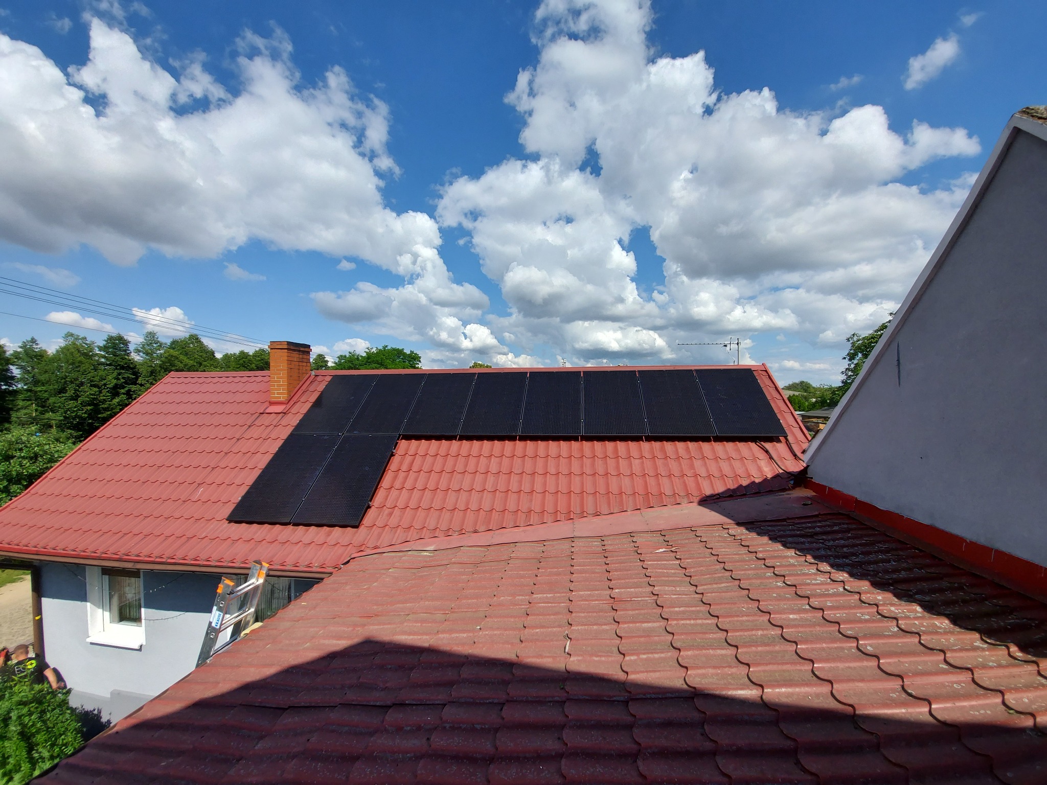 Czerwony dach z zamontowanymi panelami słonecznymi w czarnych ramach, widok z góry, błękitne niebo z białymi chmurami w tle. Drabina oparta o ścianę budynku obok.