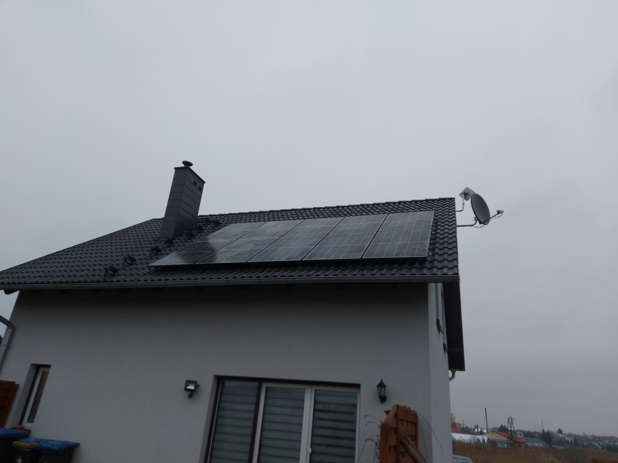 Panele słoneczne na dachu domu jednorodzinnego z kominem i anteną satelitarną w pochmurny dzień.