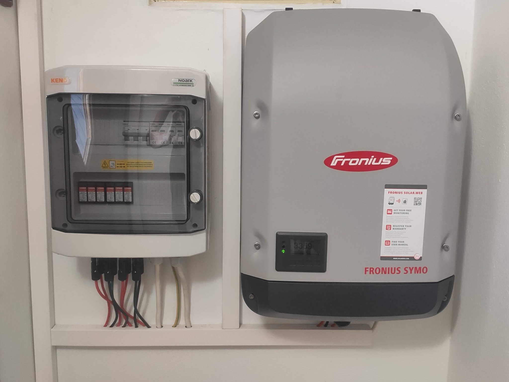 Szara skrzynka bezpiecznikowa KENO i szary inwerter Fronius Symo zamontowane na białej ścianie, widoczne przewody elektryczne.